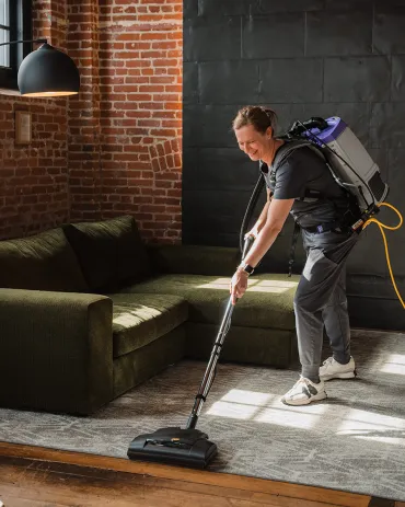 Merry Maids of Cumberland and Dauphin Counties cleaner vacuuming a living room rug with a backpack vacuum near a sectional sofa
